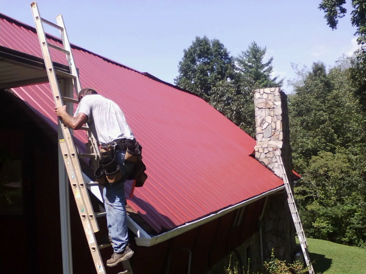 Roofer on a ladder beside a red metal roof and stone chimney — Soffit & Fascia Repair by Roofing Brothers in Menno